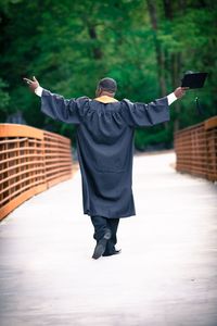 Rear view of man with arms outstretched wearing graduation gown walking on footbridge