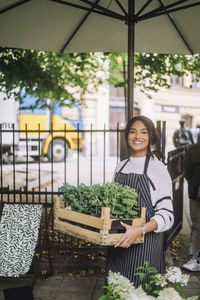 Smiling female florist holding crate of plants while standing under parasol at farmer's market