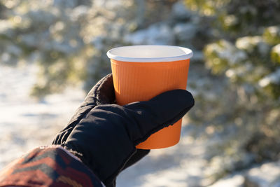 Tea mug winter travel. an orange paper cup in women's gloved hands. winter sunny blurred background