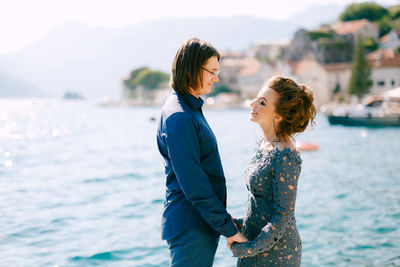 Side view of husband wife holding hands standing against sea