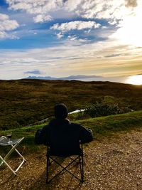 Rear view of man sitting on chair against cloudy sky