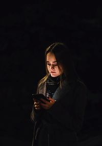 Portrait of young woman standing against black background with phone