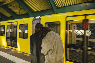 Rear view of lesbian couple standing at railroad station against yellow train