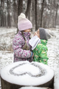 Cute sibling embracing while standing by heart shape on snow outdoors