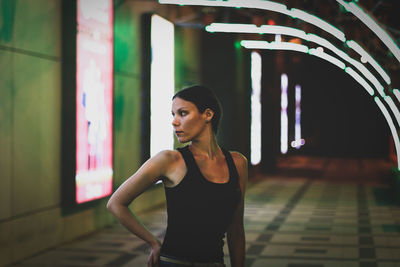 Young woman looking away while standing against wall at night