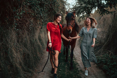 Full length of laughing women walking with horse in forest