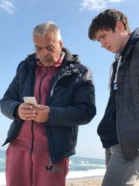 Teenage boy looking at grandfather using phone while standing at beach