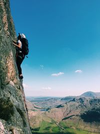 Full length of hiker climbing on rocky mountain against blue sky