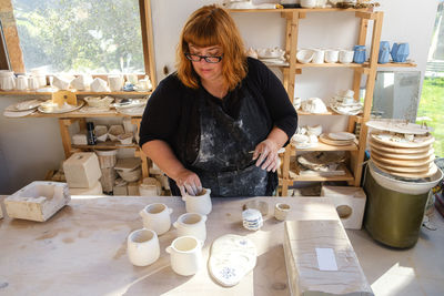 Adult female artisan in dirty apron and black clothes standing in light studio with handmade ceramic pots near clayware