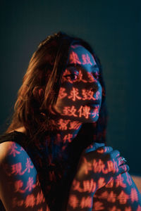 Fashionable young female model with light projection in shape of oriental hieroglyphs looking away in dark studio with red illumination