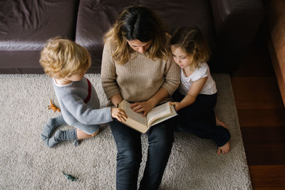 From above of young woman reading interesting fairytale in book while sitting on floor with little son and daughter at home