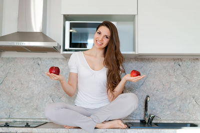 Young slim woman standing in kitchen on table, holding red apples, looking