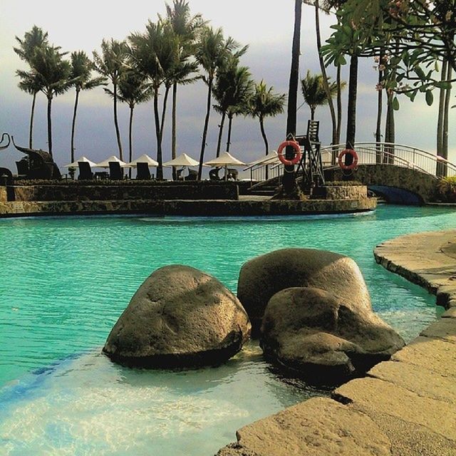 water, sea, tree, palm tree, built structure, swimming pool, blue, waterfront, architecture, rock - object, sky, building exterior, tranquility, nature, day, incidental people, rippled, beach, tranquil scene, outdoors