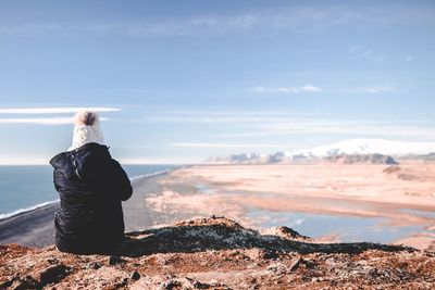 Rear view of woman looking at sea while sitting on mountain against blue sky