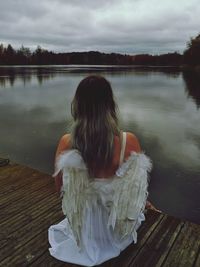 Rear view of woman wearing angel costume while sitting on pier over lake