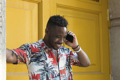 Smiling young man talking on mobile phone while standing against closed yellow doors