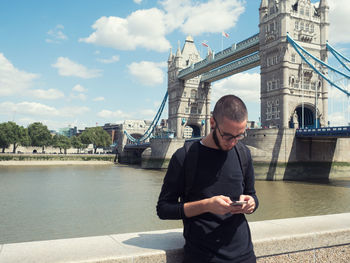 Man using mobile phone by tower bridge against sky