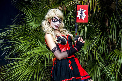 Portrait of woman in harley quinn cosplay costume holding gun with bang text against plants