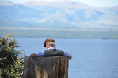 Rear view of man sitting on wooden chair by lake