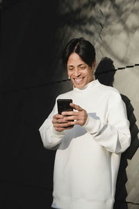 Smiling asian man in white jumper standing on street concrete wall and reading messages on smartphone