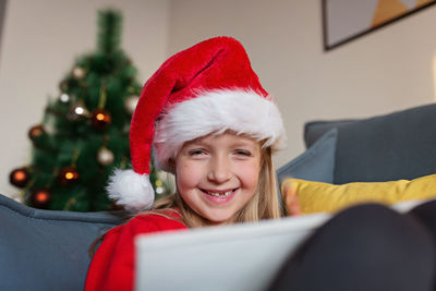 Cute blonde caucasian girl 7 years old sitting on couch near christmas tree writing letter to santa