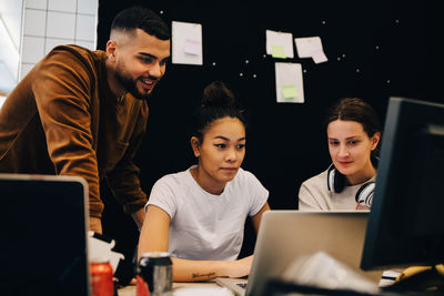 Colleagues looking at businesswoman using laptop against bulletin board at creative office