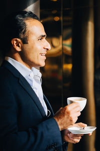 Side view of smiling mature businessman holding coffee cup while looking away at cafe