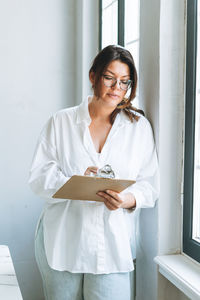 Young woman plus size in white shirt near window with pen and documents in hands in the office