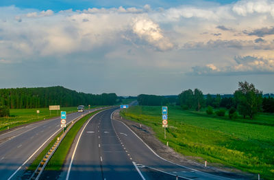 View of a summer paved highway with road signs among hills and a green field, along which cars