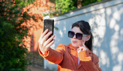 A young brunette girl in an orange jacket takes a selfie against a brick wall