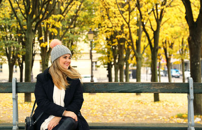 Smiling young woman sitting on bench in park during autumn