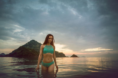 Woman in bikini standing in sea against cloudy sky during sunset