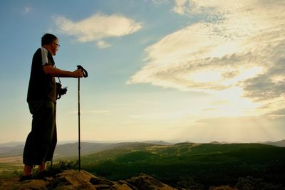 Photographer with camera on neck and poles in hands on peak. dreamy valley below rocky mountains