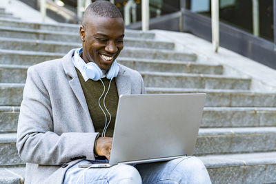 Smiling businessman with headphones using laptop on steps