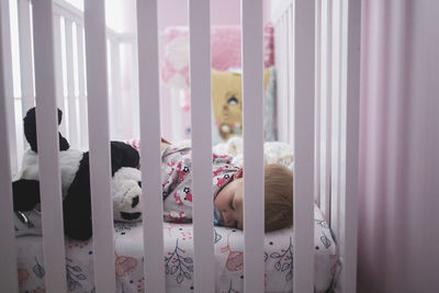 Baby girl sleeping with stuffed toy in crib seen through railings at home