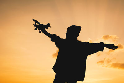 Silhouette girl holding toy airplane against sky during sunset