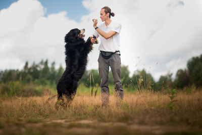 Man playing with dog while standing on grassy land against cloudy sky