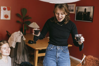 Cheerful woman holding drink glasses while celebrating with female friend in apartment