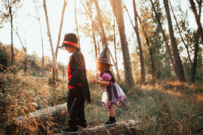 Side view full length of girl wearing witch hat standing with brother at forest during halloween