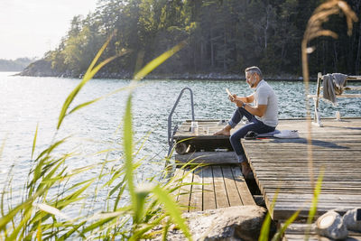 Full length of man using digital tablet while sitting on wooden pier over lake at forest