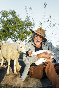 Happy farmer carrying goose sitting by sheep at farm on sunny day