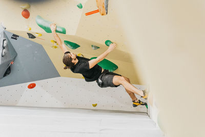 Full body side view of sportive male alpinist climbing wall with colorful grips during intense training in light modern gym