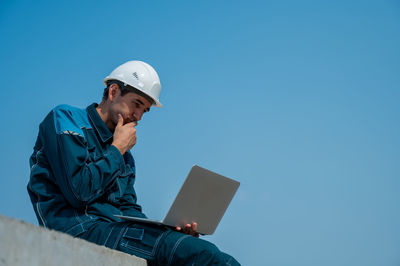 Side view of man using laptop while standing against clear blue sky