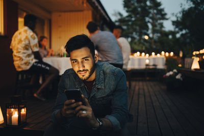 Portrait of smiling man holding smart phone while friends in background during dinner party