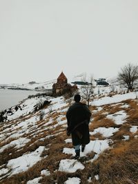 Rear view of man walking towards old church on snow covered hill by lake against clear sky