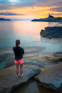 Rear view full length of woman standing on rock looking at sea during sunset