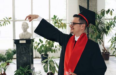 Optical illusion of smiling mid adult man in graduation gown holding statue