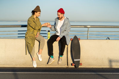 Happy couple drink hot tea outdoors sit on bridge wear trendy street clothes with longboard near