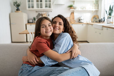 Happy family single mother and teenage daughter best friends hugging while sitting on sofa at home