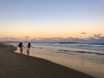 Sisters running at beach against sky during sunset